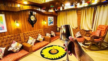 Une pièce avec un plafond orné de motifs, un ensemble de canapés, une table décorée d’un motif solaire et une main tenant un verre.