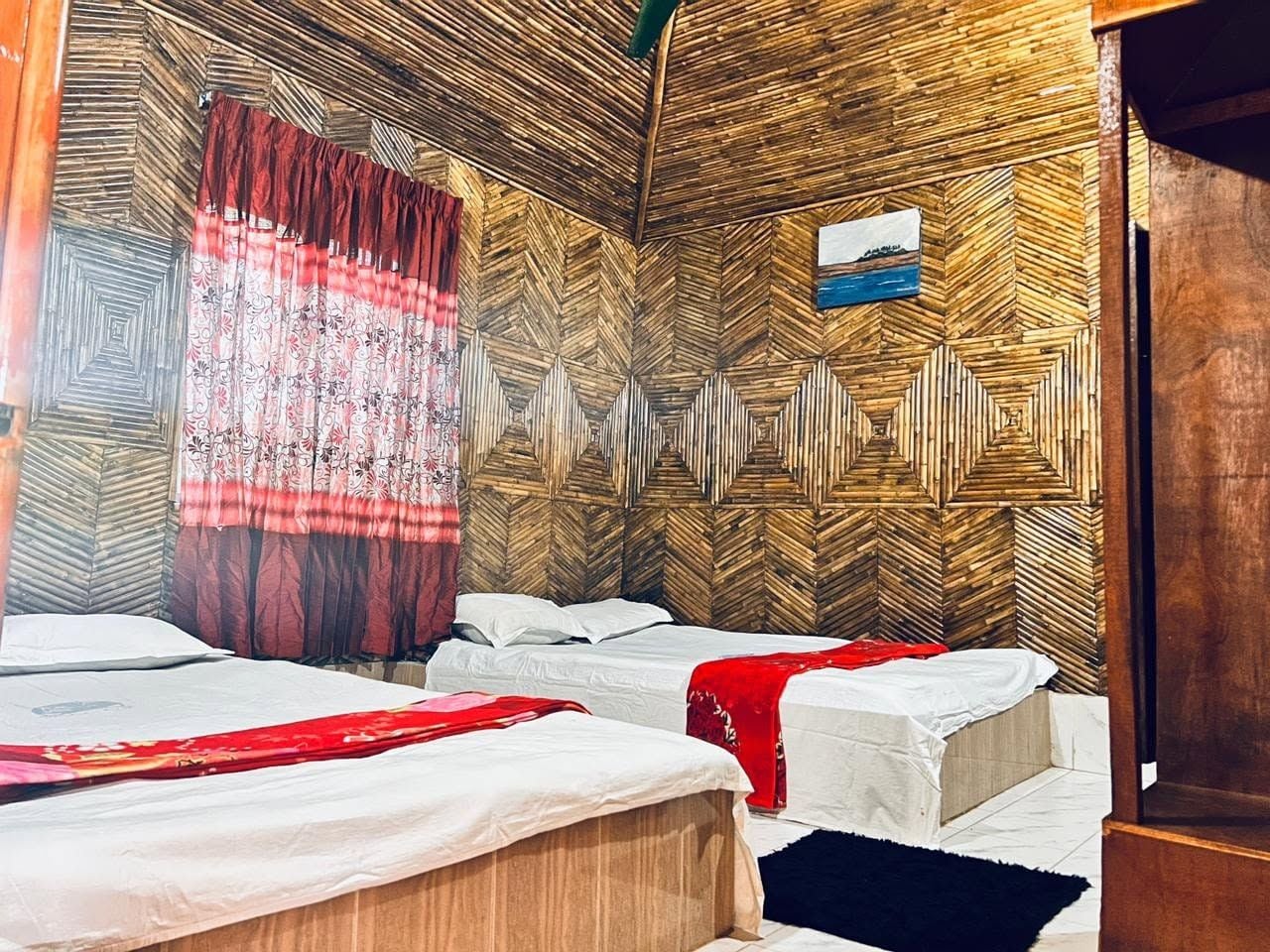 Une chambre avec deux lits, un mur en bois orné d’un motif géométrique, un rideau rouge et un petit tableau encadré accroché au mur.