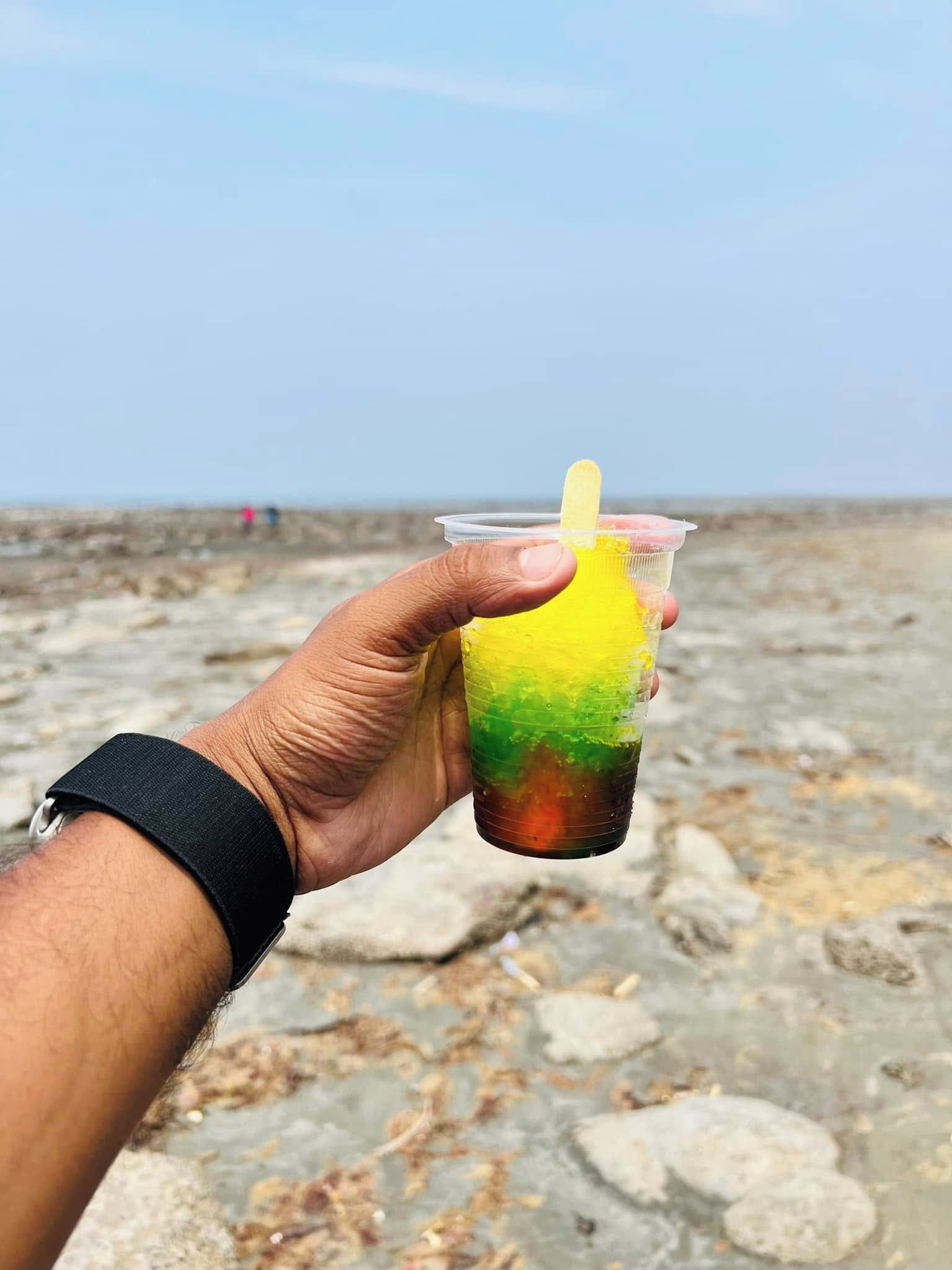 A hand holding a plastic cup with a straw, containing a layered drink with yellow, green, and brown colors, against a rocky beach background.