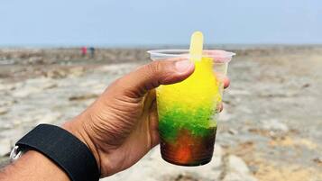 A hand holding a plastic cup with a straw, containing a layered drink with yellow, green, and brown colors, against a rocky beach background.