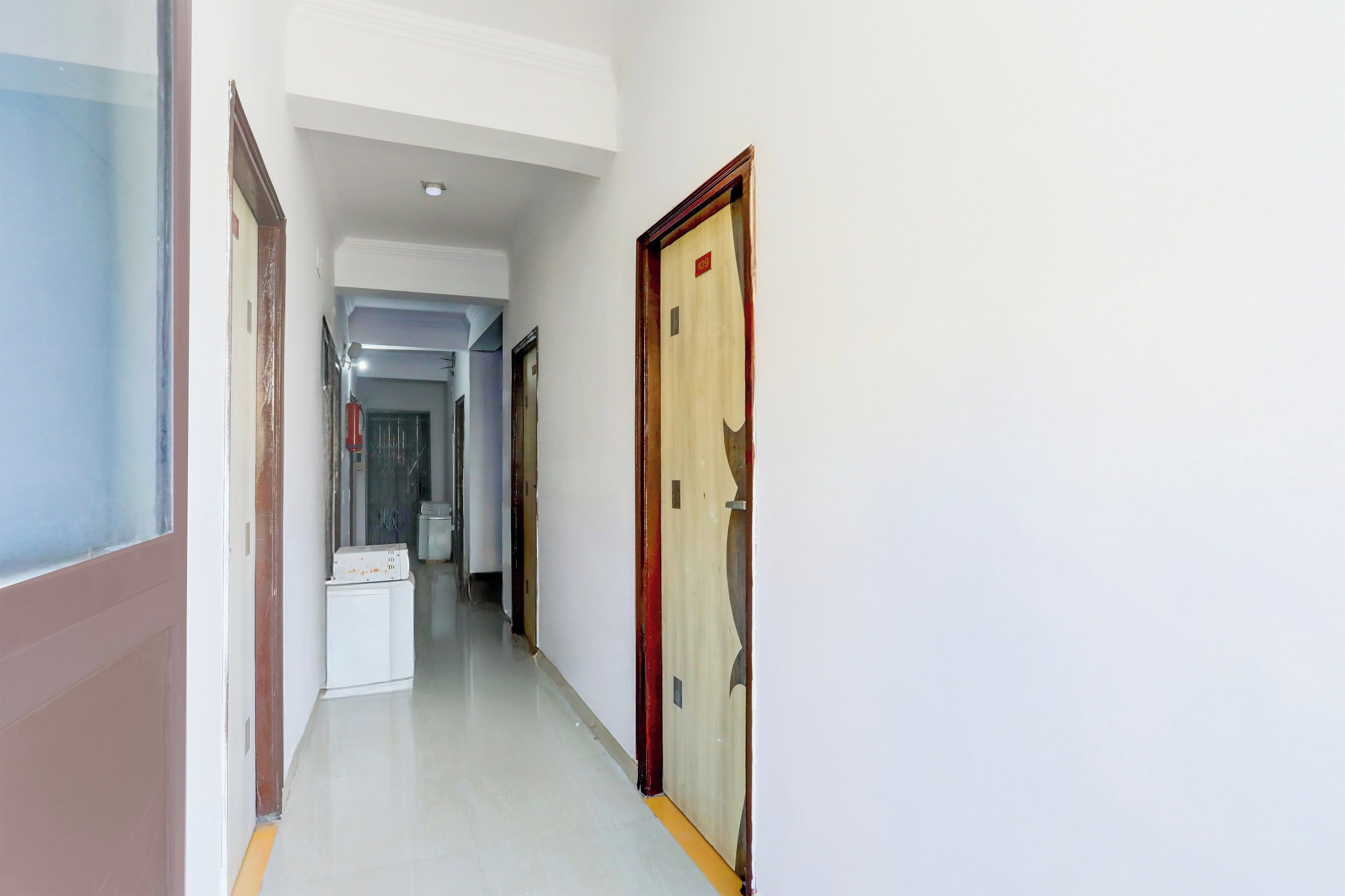 Un pasillo con pared blanca, una puerta de marco rojo y un electrodoméstico blanco sobre un pedestal.