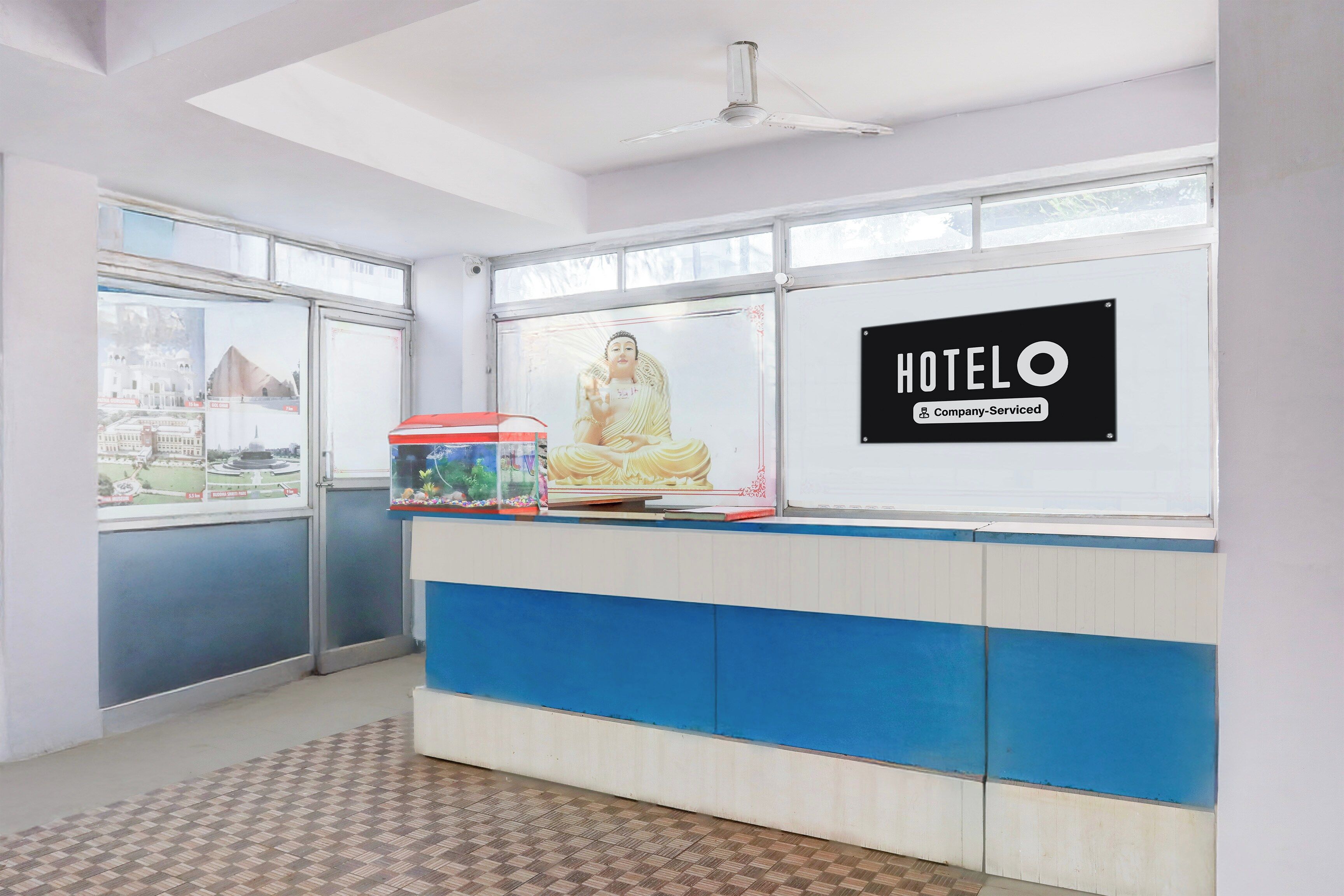 Balcão de recepção de hotel com uma grande estátua de Buda, um cartaz emoldurado com vista da cidade e uma placa indicando que o hotel oferece serviços corporativos.
