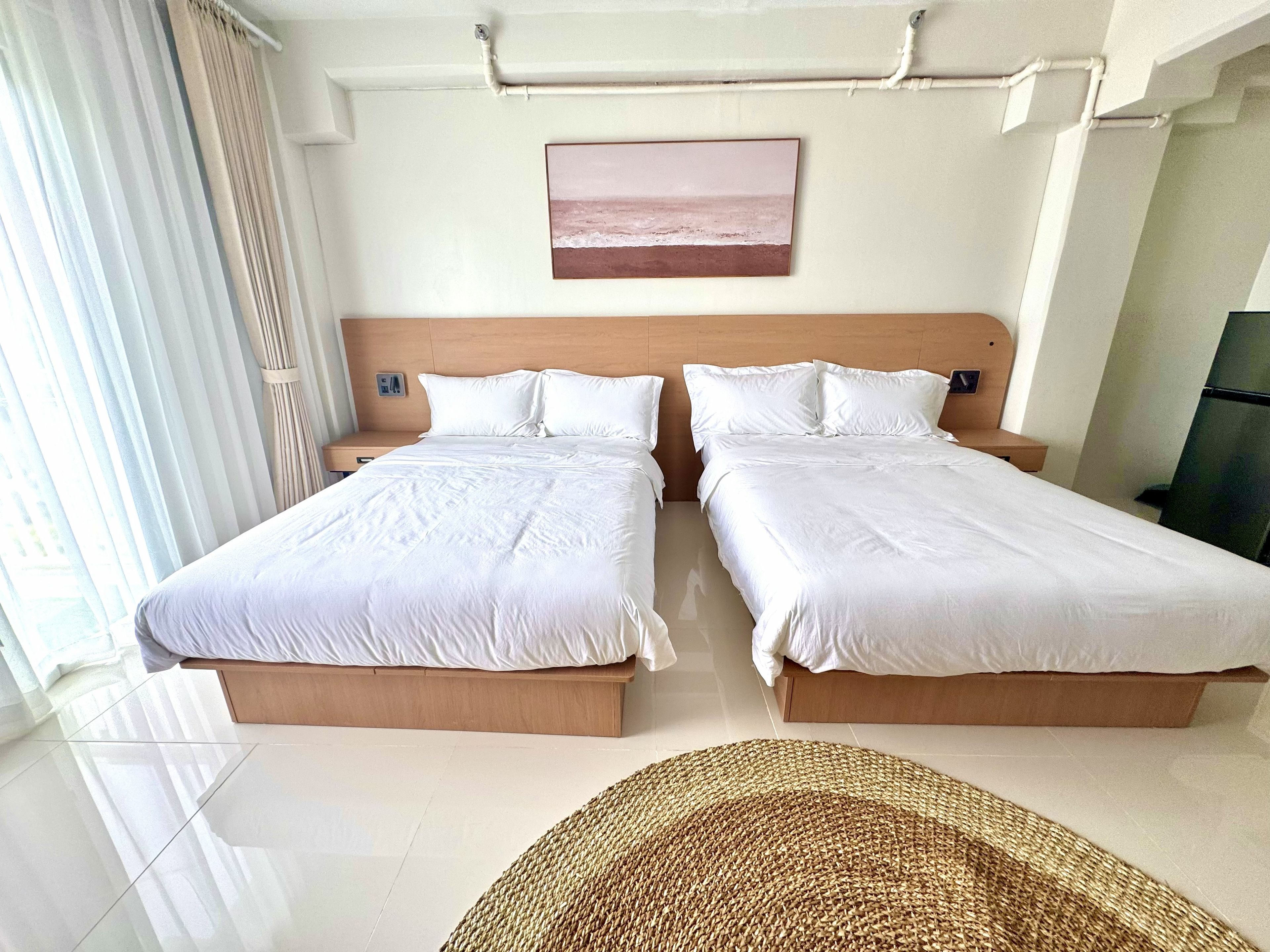 Habitación de hotel con dos camas, un ventanal con cortinas, un cuadro colgado en la pared y una alfombra tejida en el suelo.