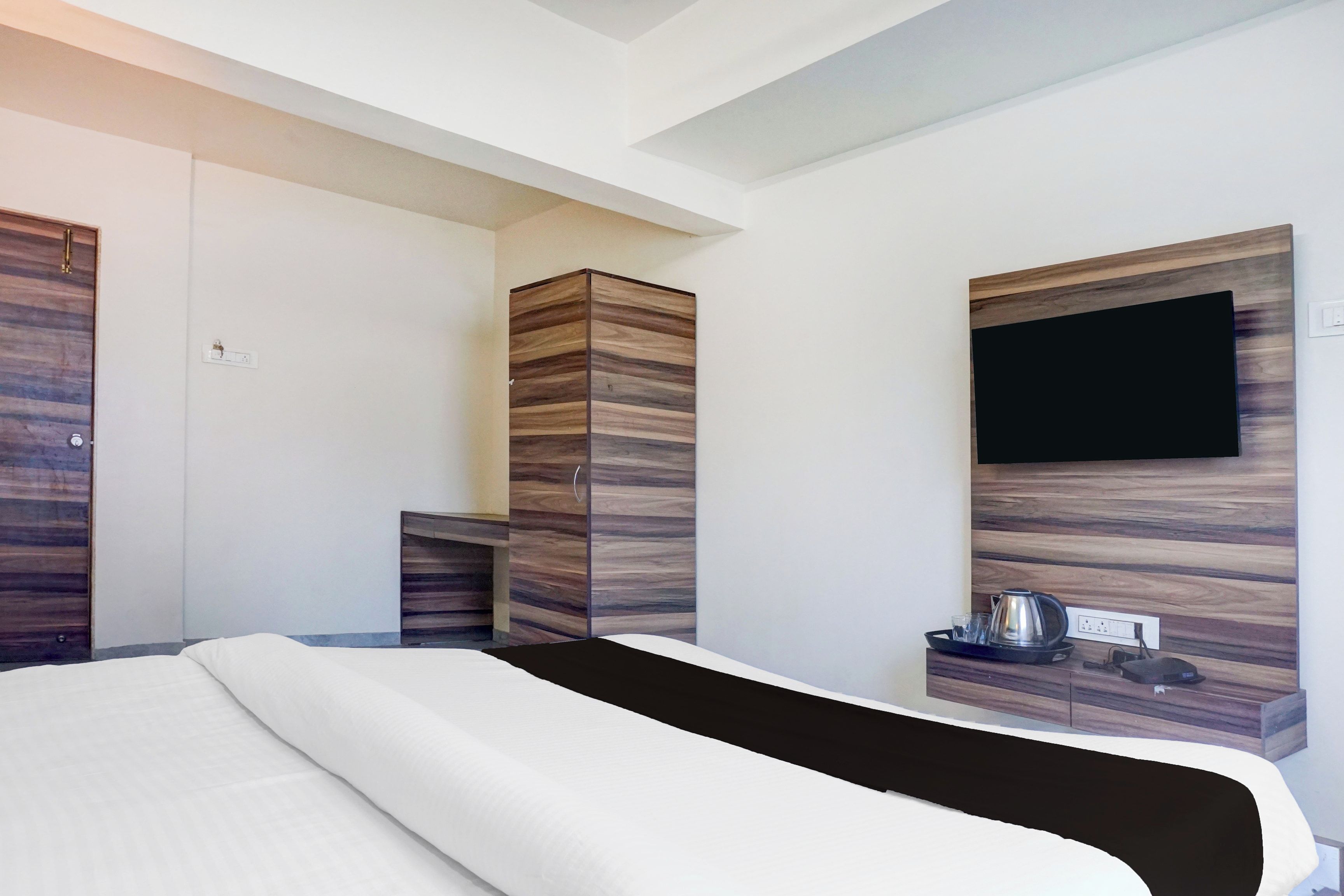 A hotel room with a bed, a wardrobe, a television mounted on the wall, and a small table with a kettle and glasses.