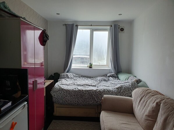 Ein gemütlicher Raum mit Bett, Schreibtisch, Küchenzeile und einem Fenster mit Vorhängen.