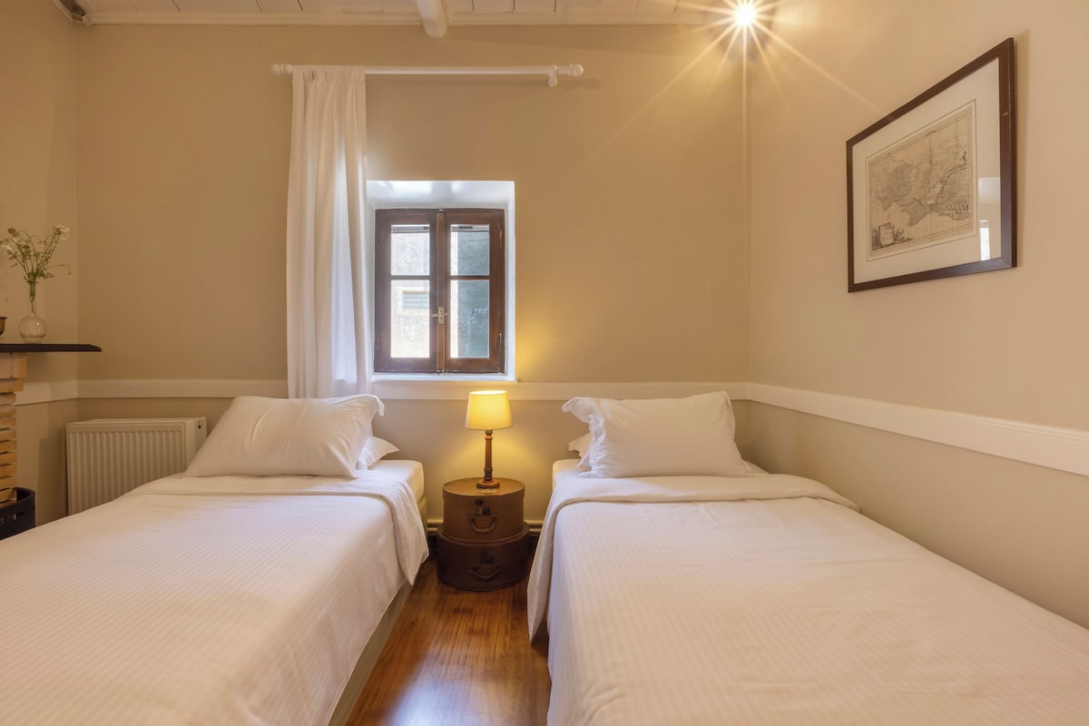 A room with two single beds, a window with curtains, a bedside lamp, a radiator, and a framed map on the wall.