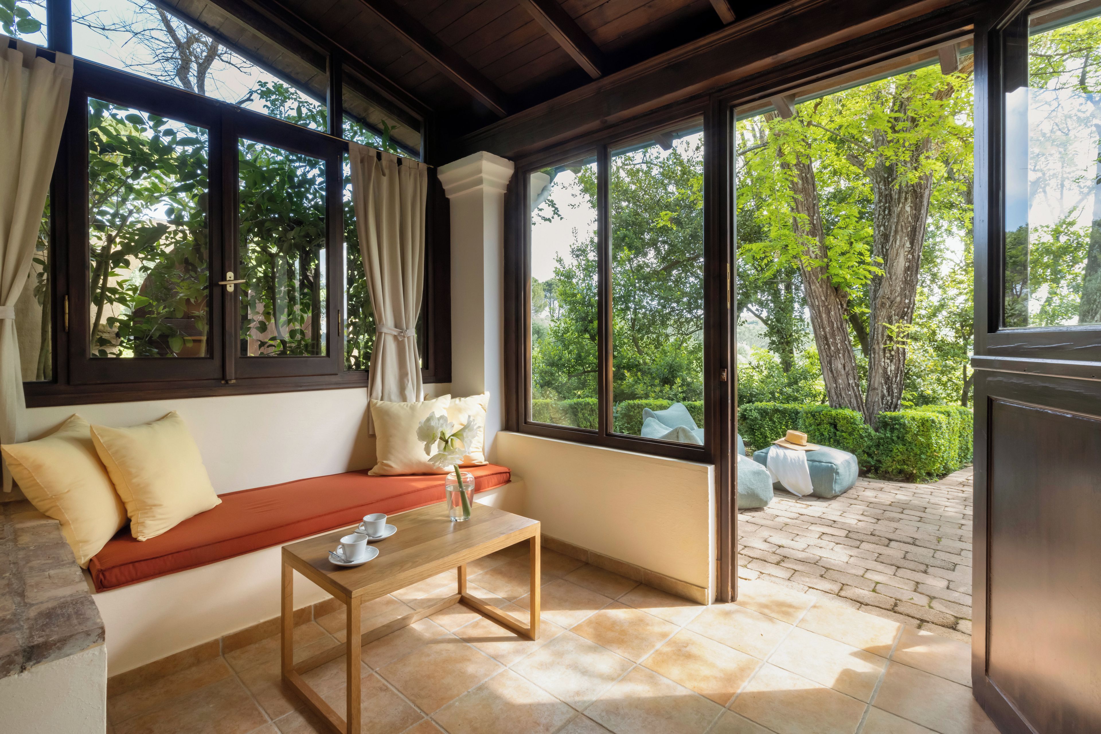 Una habitación con un banco de madera, una mesita y vistas a un jardín a través de amplios ventanales.