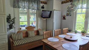 A cozy room with a wooden sofa, a dining table with placemats, a television mounted on the wall, and a window with curtains.