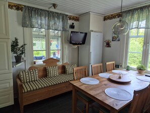 A cozy room with a wooden sofa, a dining table with placemats, a television mounted on the wall, and a window with curtains.