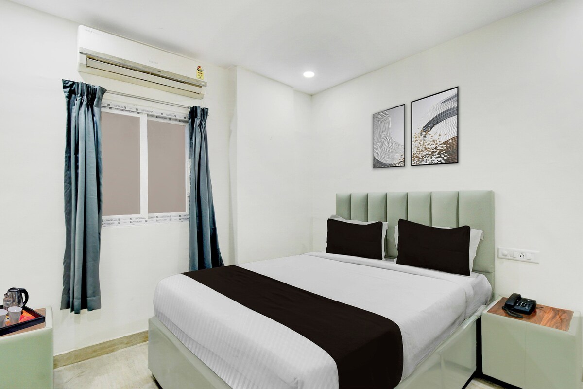 A hotel room with a bed, a bedside table, a nightstand, a window with curtains, and wall art.