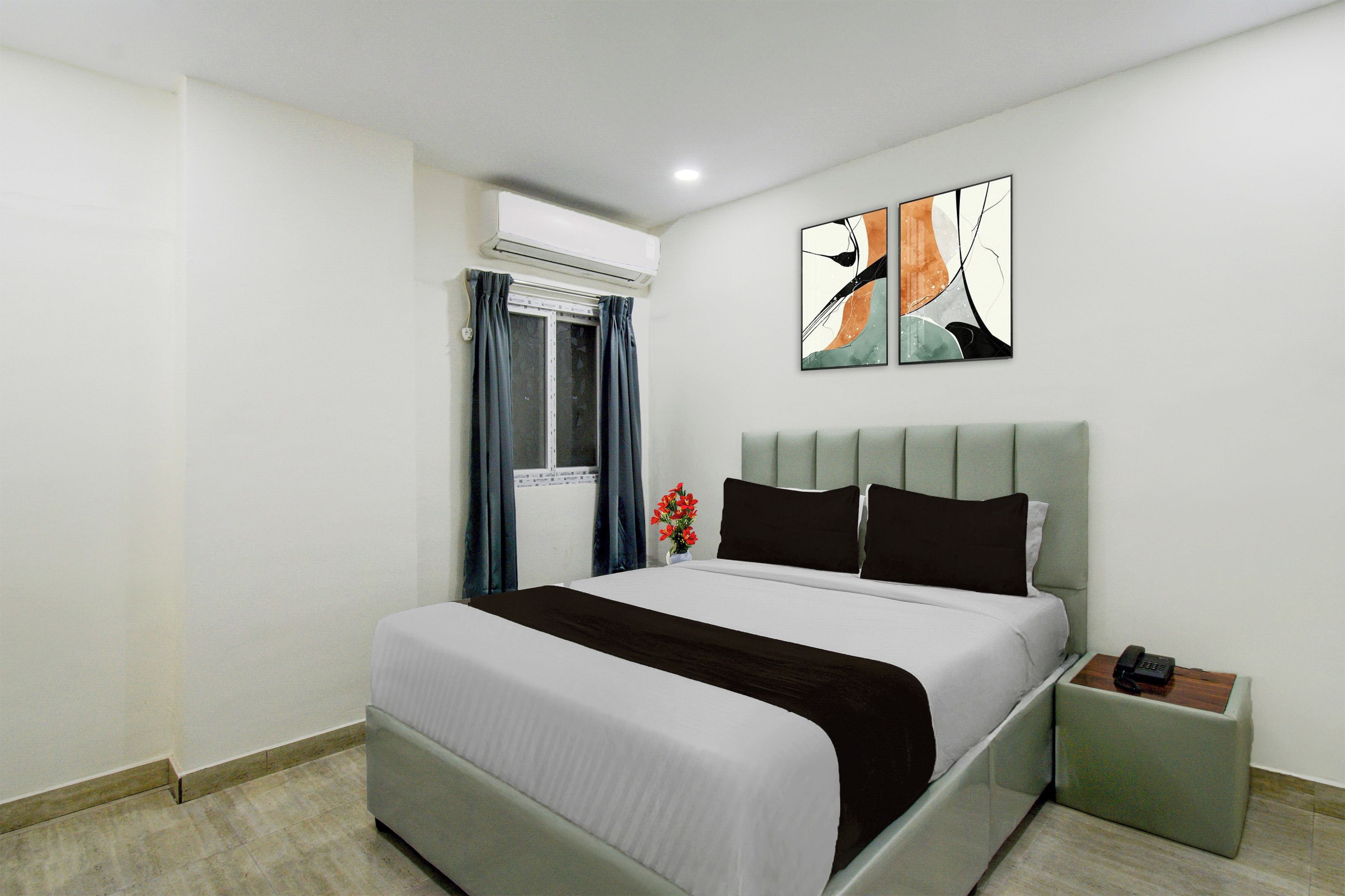 A modern hotel room with a bed, a bedside table, a nightstand, an air conditioning unit, a window with curtains, and a wall-mounted abstract art piece.
