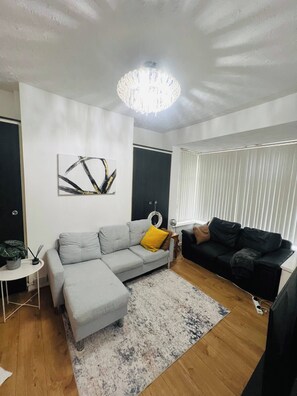 Uma sala de estar com um sofá cinzento, um sofá preto, uma mesinha branca, um abajur e um quadro na parede.