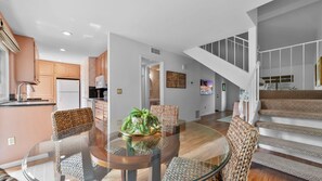Ein modernes Interieur mit einem Essbereich, der einen Glastisch und gewebte Stühle umfasst, einer Küche mit Holzschränken und einer Treppe, die zu einer oberen Ebene führt.