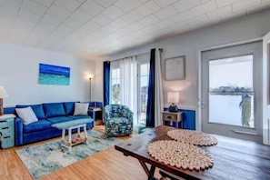 A living room with a blue sofa, a patterned armchair, a wooden coffee table, and a view of a body of water through large windows.