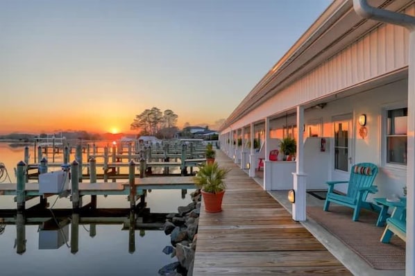 A waterfront hotel with a wooden deck, outdoor seating, and a view of the sunset over the water.