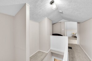 A narrow hallway with white walls and a textured ceiling, leading to a room with a bed and a small table. - Sunnyside Gameday House (South Bend)