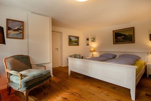 Ein Schlafzimmer mit Holzboden, einem weißen Bett, einem Sessel und an den Wänden befestigten Bildern.