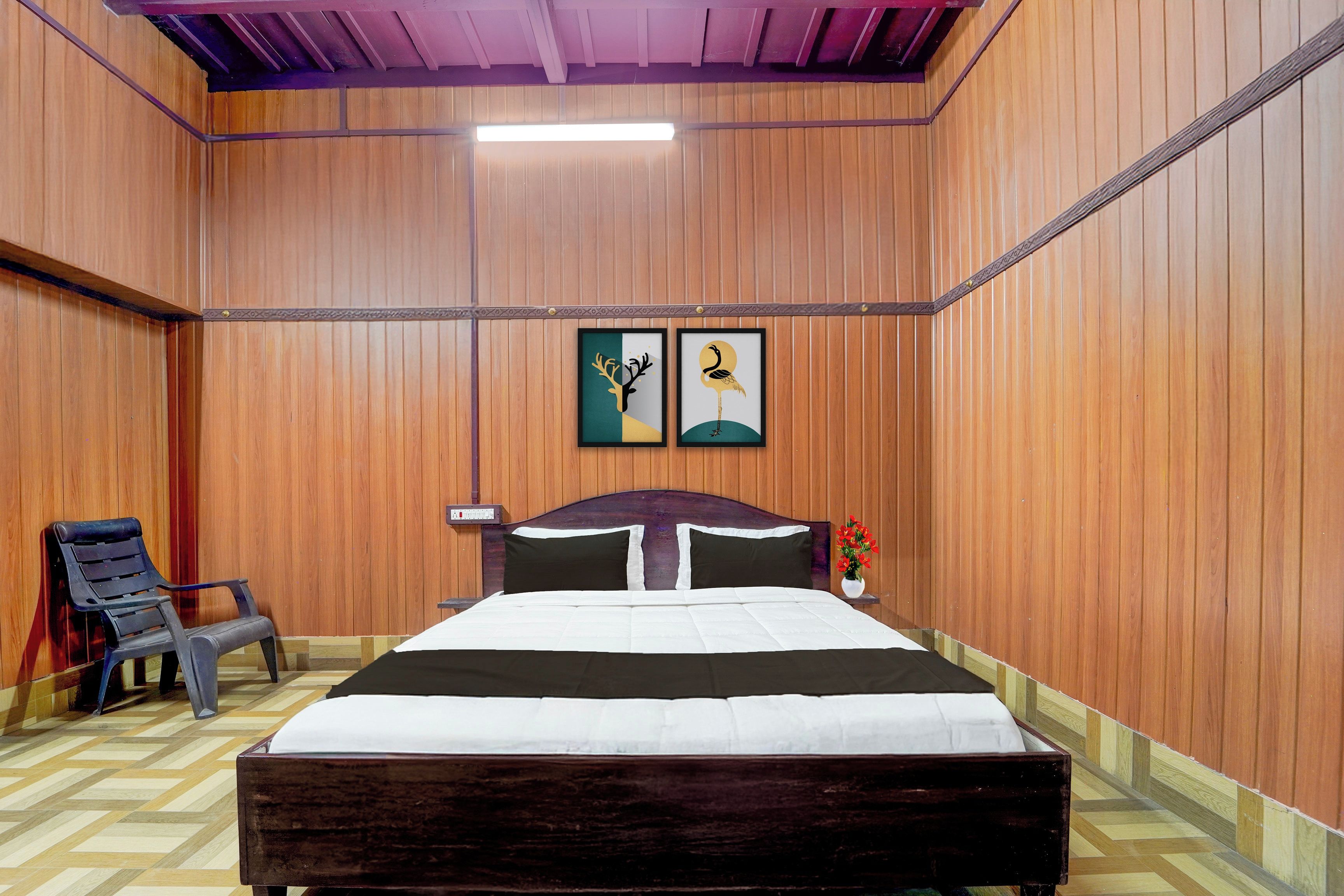 Una habitación con paneles de madera, una cama con cabecera oscura, dos cuadros en la pared, una silla y una mesita con un florero.