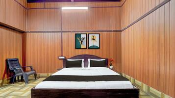 Una habitación con paneles de madera, una cama con cabecera oscura, dos cuadros en la pared, una silla y una mesita con un florero.