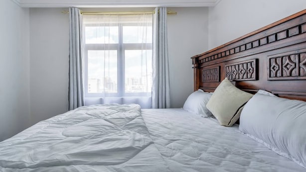 Un lit bien fait, avec du linge de lit blanc, dans une pièce dotée d’une grande fenêtre et d’une tête de lit en bois.