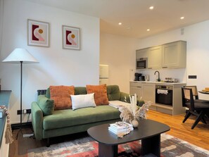 Ein modernes Wohnzimmer mit einem grünen Sofa, einem Couchtisch mit Büchern und einer Küchenzeile inklusive Mikrowelle und Backofen.