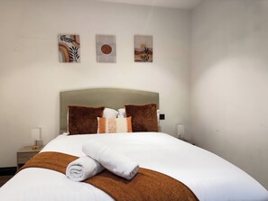 Ein ordentlich bezogenes Bett mit einer braun-weißen Decke, zwei Kissen und einem zusammengerollten Handtuch. Darüber hängen drei Bilder an der Wand.