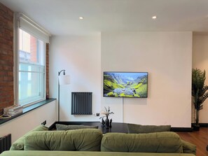 Ein modernes Wohnzimmer mit einem grünen Sofa, einem flachbildfernseher an der Wand, einer Heizung und einem Fenster mit Blick auf ein Gebäude.