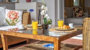Una disposición para el desayuno con una mesa de vidrio, una silla de mimbre, un sofá y una planta en maceta.
