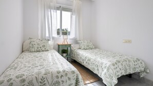 Un dormitorio con dos camas individuales, una mesita de noche pequeña con una lámpara, una ventana con cortinas translúcidas y vistas a zonas verdes.