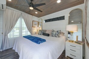 Ein Schlafzimmer mit einem Bett, einem Deckenventilator, einem Nachttisch mit Lampe, einem Spiegel und einem Fenster mit Vorhängen.