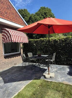 Eine Terrasse mit Tisch und Stühlen, einem roten Sonnenschirm und einem Ziegelgebäude mit gestreiftem Markisenvorbau.