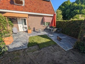 Ein Haus mit Ziegelaußenwand, einem Ziegeldach, einer überdachten Terrasse mit Gartenm öbeln und einem Hund, der auf der Terrasse spazieren geht.
