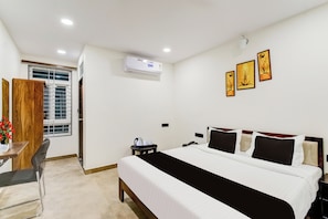 A hotel room with a bed, bedside table, chair, desk, and a window with blinds. - Townhouse OAK Owaisi Hospital (Hyderabad)