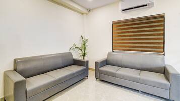 A waiting area with two grey sofas, a potted plant, a window with blinds, and an air conditioning unit on the wall.
