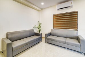 A waiting area with two grey sofas, a potted plant, a window with blinds, and an air conditioning unit on the wall. - Townhouse OAK Owaisi Hospital (Hyderabad)