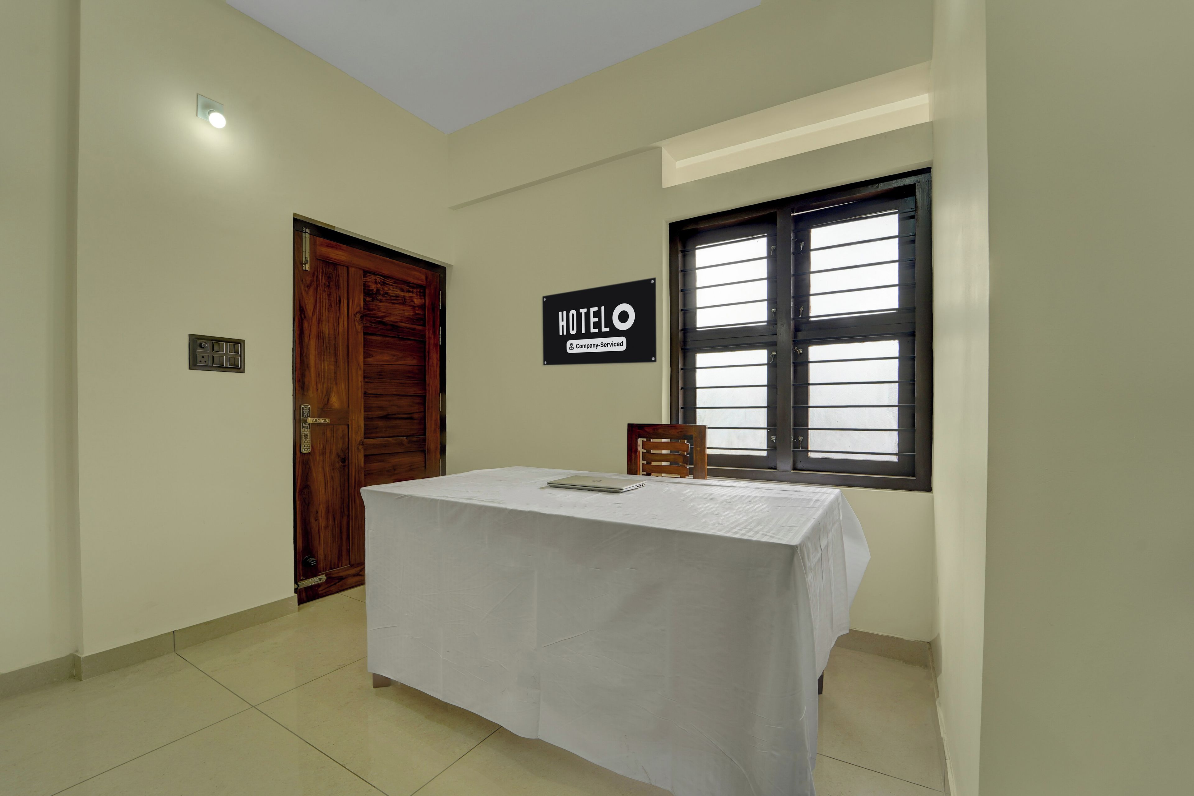 Une chambre d’hôtel avec une porte en bois, une pancarte indiquant qu’elle est mise à disposition par l’entreprise, une fenêtre avec des stores et une table sur laquelle se trouve un ordinateur portable.
