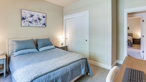 Un dormitorio con cama, mesitas de noche, lámpara, puerta y un cuadro en la pared.