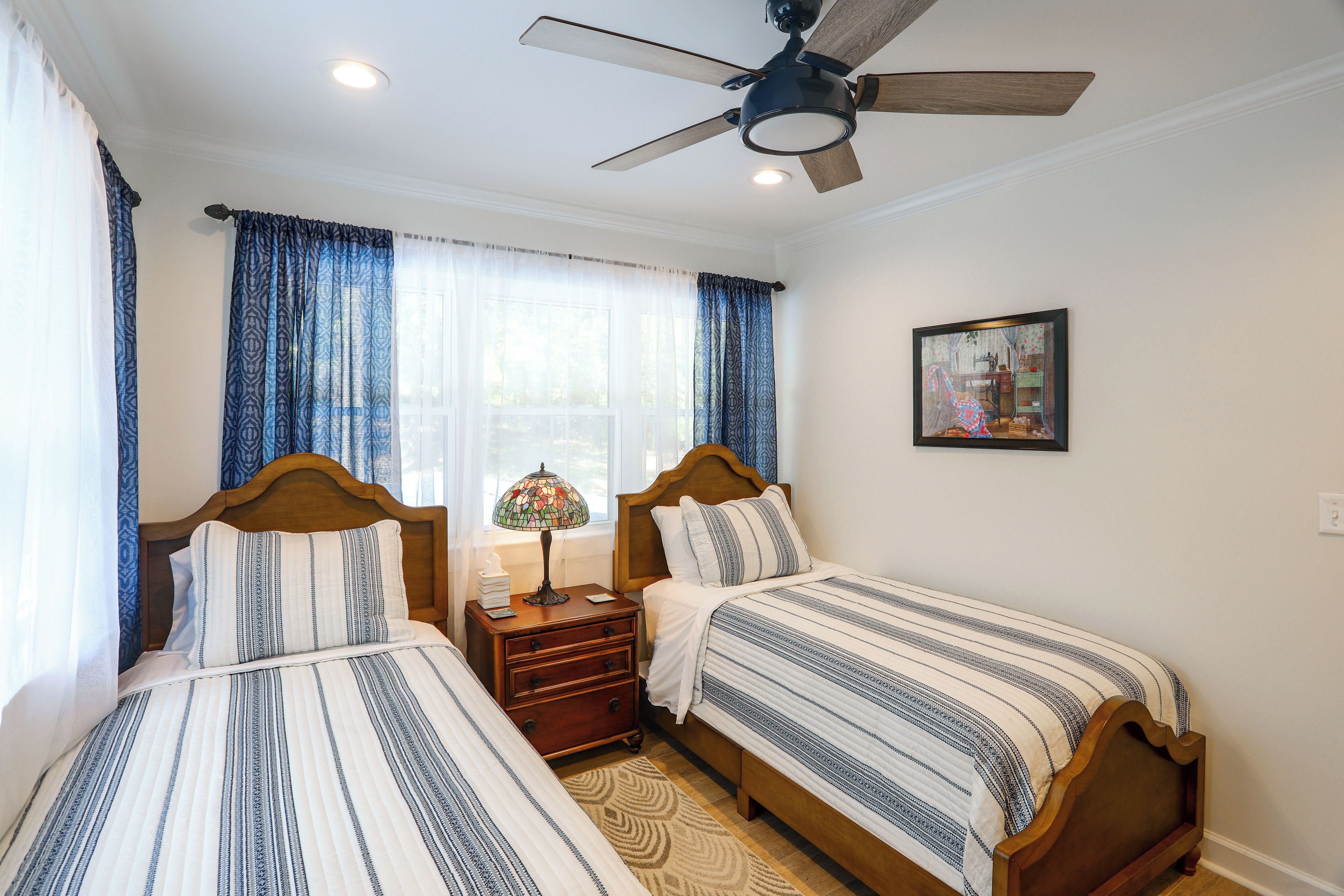 Quarto de hotel com duas camas de solteiro, um ventilador de teto, mesa de cabeceira com abajur e um quadro na parede.