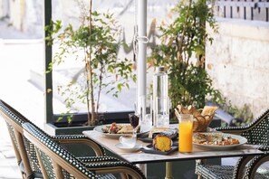 Et udendørs spisebord dækket med forskellig mad og drikke, omgivet af fletstole og grønne planter.