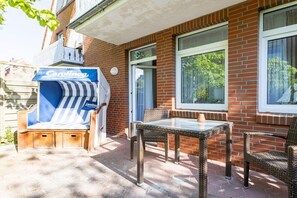 Eine Terrasse mit einem Korbstuhl-Tisch und -Stühlen, einem blau-weiß gestreiften Liegestuhl und einem Fenster mit Blick auf Grünflächen.