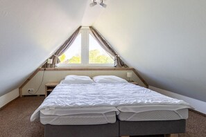 A bedroom with a sloped ceiling, a bed with white bedding, a nightstand, and a window with curtains. - Sielwärts - Spiekeroog (Carolinensiel)
