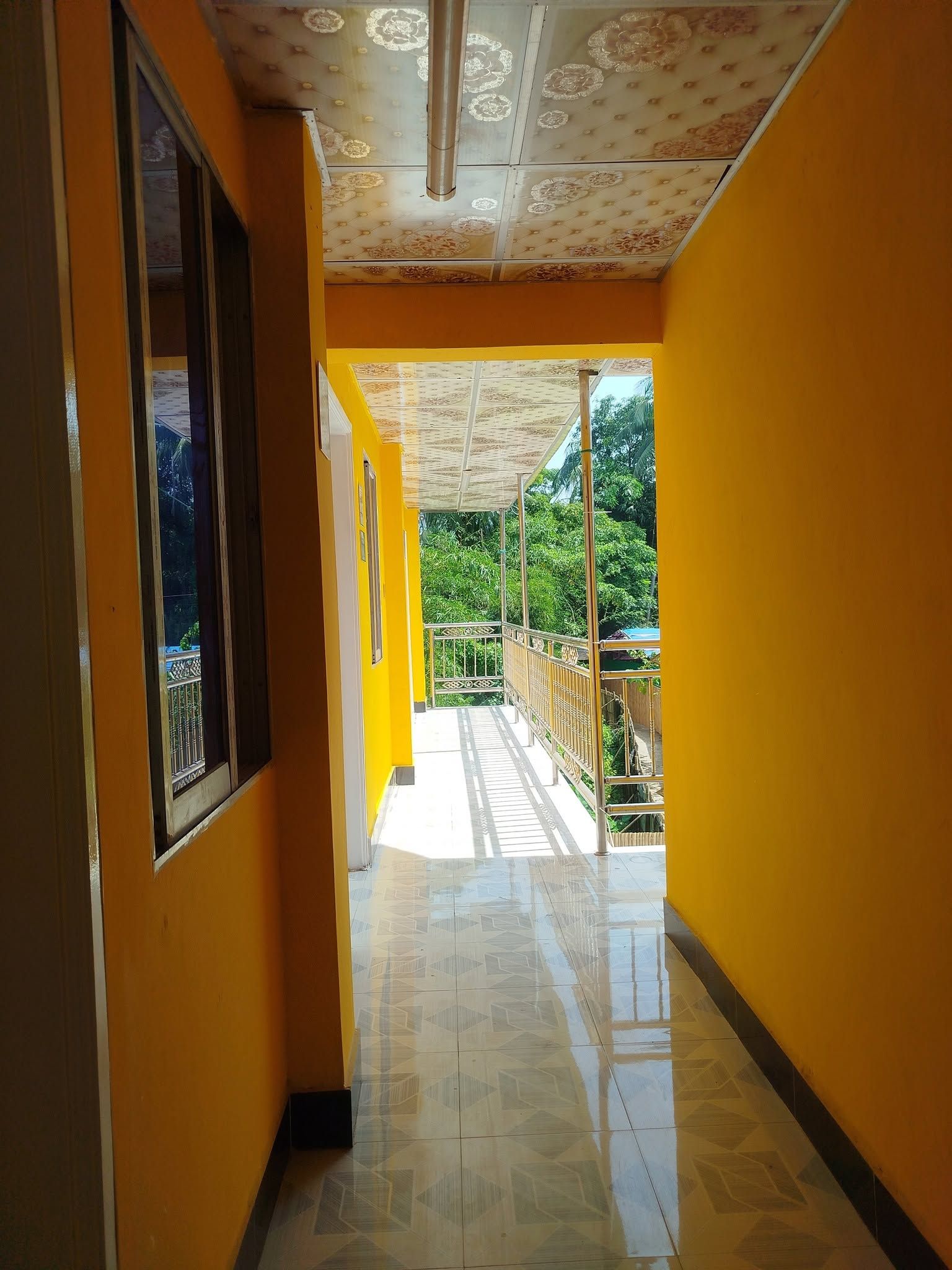 Un couloir aux murs jaunes, doté d’un sol carrelé et d’un balcon avec une rambarde en métal.