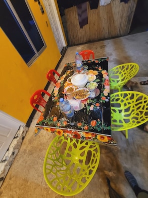 A dining table with floral tablecloth, surrounded by chairs, in a room with yellow walls and a concrete floor.