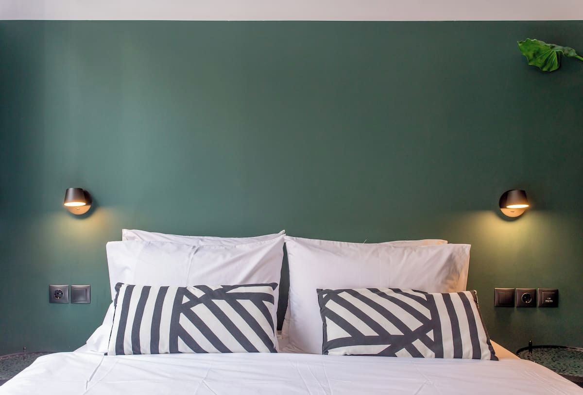 Una cama con ropa de cama blanca y almohadas a rayas negras y blancas, dos lámparas de pared y una pared verde.