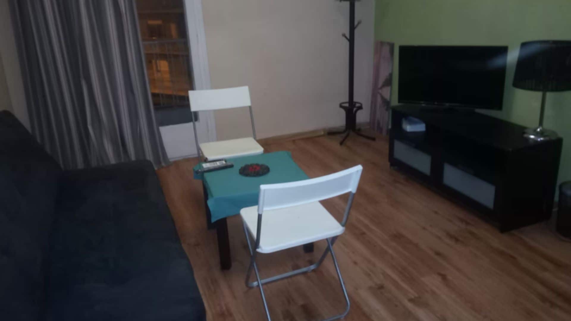 Una sala de estar con un sofá negro, una mesa de centro verde azulado, una silla plegable blanca, un mueble de televisión con televisor y una lámpara de pie.