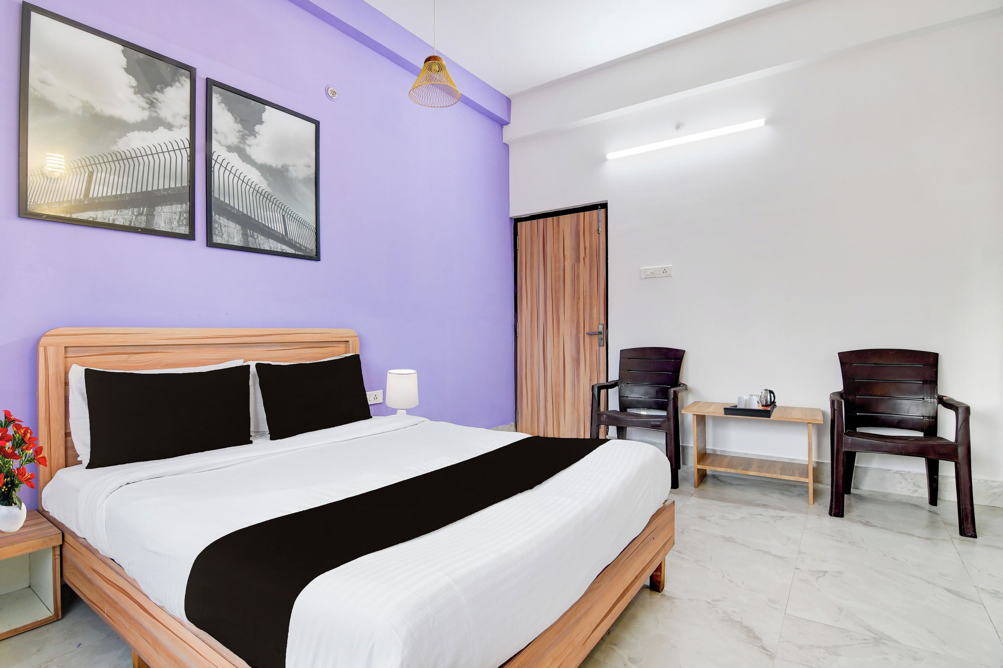 Quarto de hotel com uma cama, uma escrivaninha, duas cadeiras, um abajur e duas imagens emolduradas na parede.