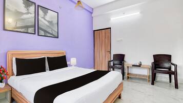 Quarto de hotel com uma cama, uma escrivaninha, duas cadeiras, um abajur e duas imagens emolduradas na parede.