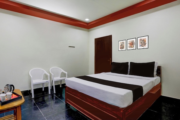 A hotel room with a bed, two chairs, a small table with a kettle and cups, and three framed pictures on the wall.