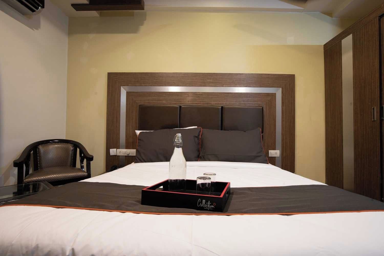 A hotel room with a large bed, a wooden headboard, a bedside table with a bottle and glasses, and a chair.