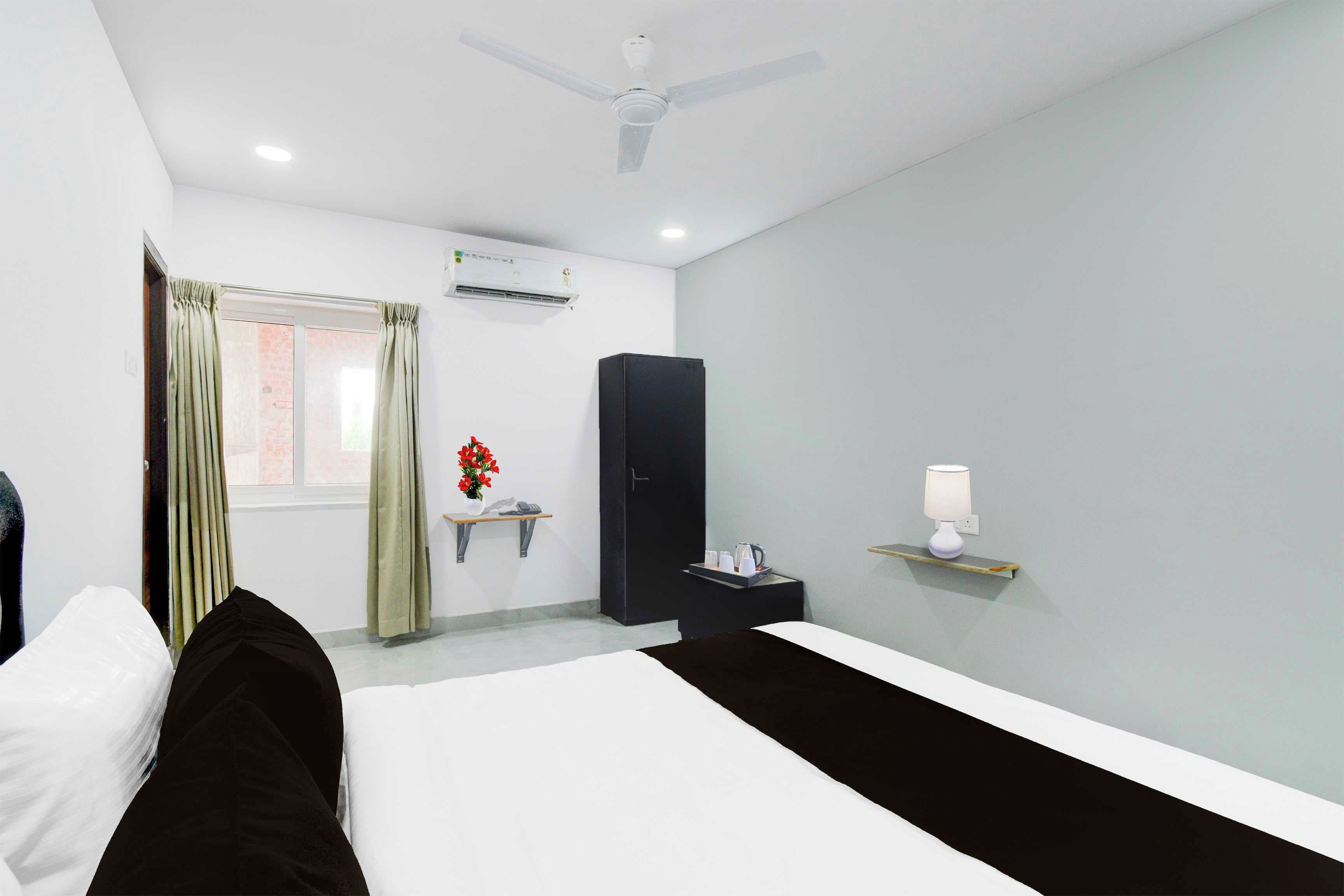 A hotel room with a bed, a bedside table with a lamp, a wardrobe, an air conditioning unit, and a window with curtains.